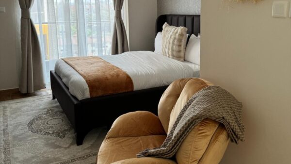 Cozy bedroom with a bed, brown and white bedding, velvet chair with a throw, and large windows with sheer curtains.