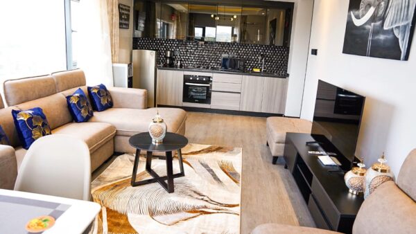 Modern living room with beige sofas, blue floral pillows, black coffee table, patterned rug, TV, and kitchen in the background.