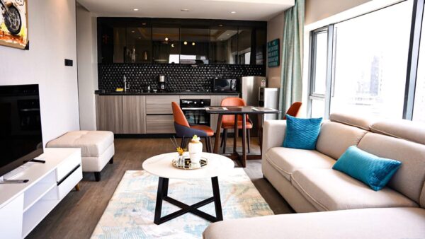Modern living room with beige sofa, blue cushions, round coffee table, and open kitchen with black backsplash and dining set.
