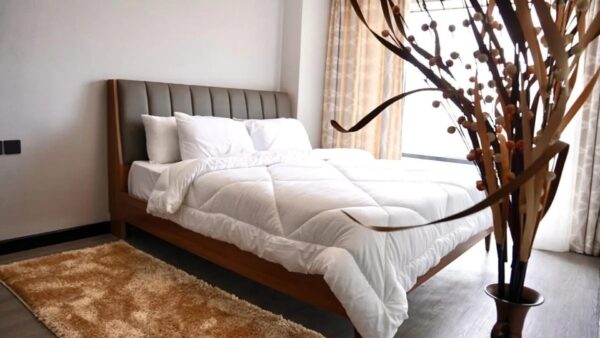 Minimalist bedroom with a neatly made bed, brown headboard, white bedding, beige rug, and a tall vase with decorative branches.