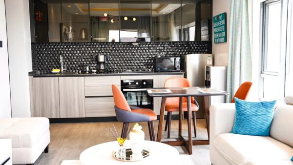Modern kitchen with wood cabinets, black backsplash, dining table, orange chairs, and a cozy living area with a blue pillow.