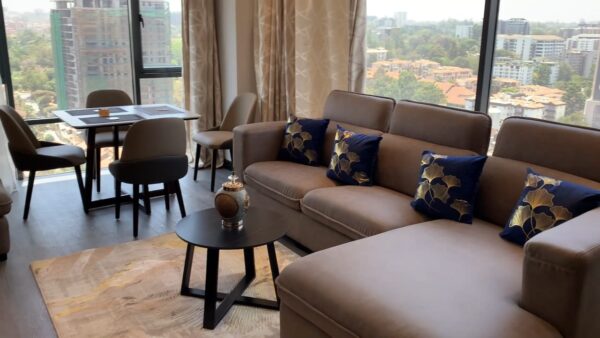 Modern living room with beige sofa, blue patterned pillows, glass windows with city view, dining table, and decorative vase.
