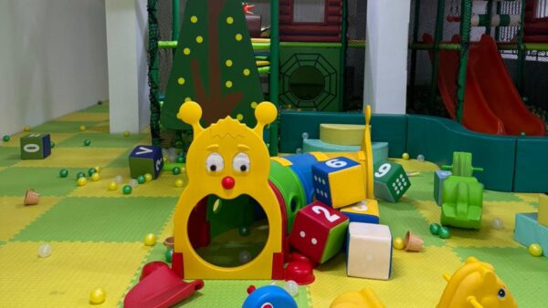 Colorful indoor playroom with foam mats, toy blocks, a slide, and a caterpillar tunnel surrounded by scattered balls.
