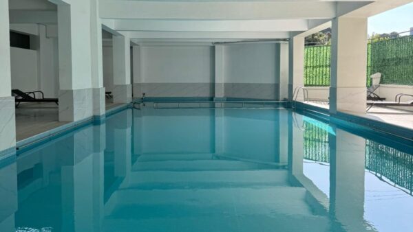 Indoor swimming pool with clear blue water, surrounded by white pillars and sun loungers visible on the tiled deck.