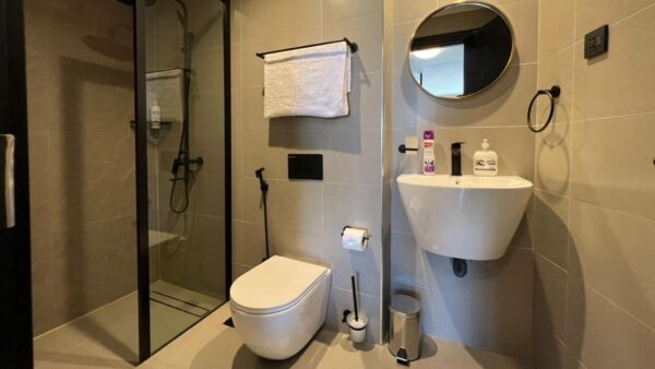 Modern bathroom with glass shower, wall-mounted toilet, round mirror above white sink, towel rail, and soap dispenser.