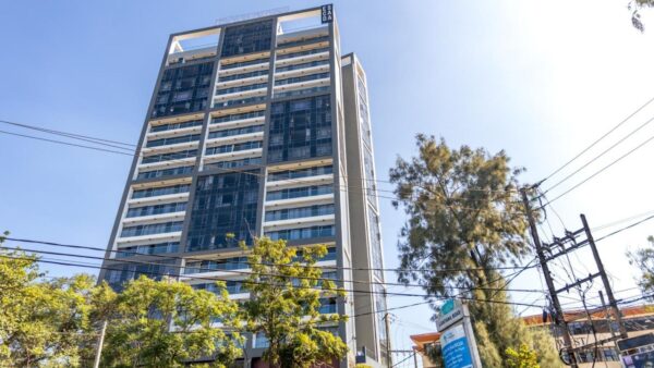 Tall modern building with glass facade, blue sky background, surrounded by trees and power lines.
