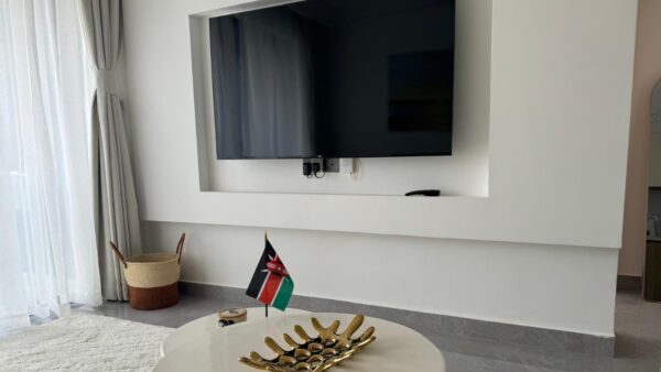 Wall-mounted TV above a shelf, small Kenyan flag, gold decorative tray on round table, white curtains, woven basket.
