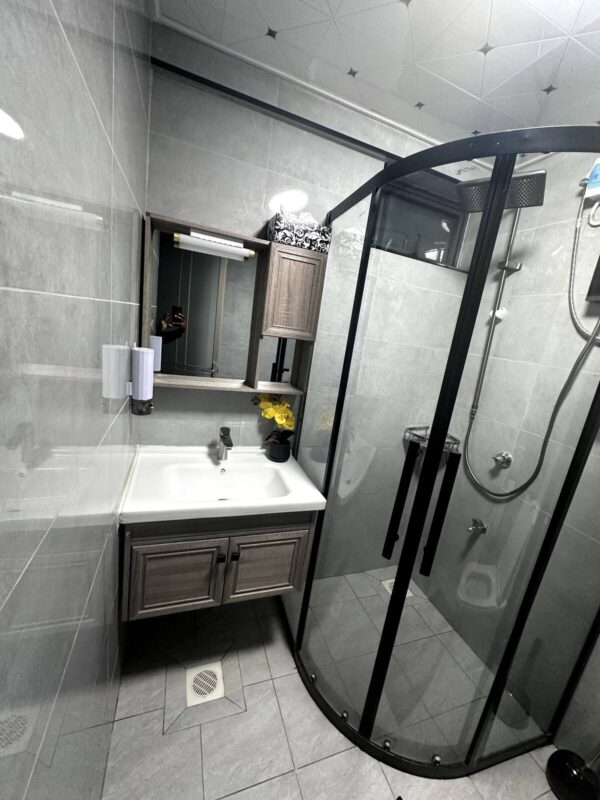 Modern bathroom with glass shower, gray tiles, wooden vanity, mirror, and yellow flowers on the sink.