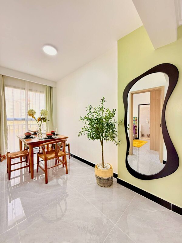 Dining area with wooden table set for four, large plant, wavy mirror on green wall, and bright window light.