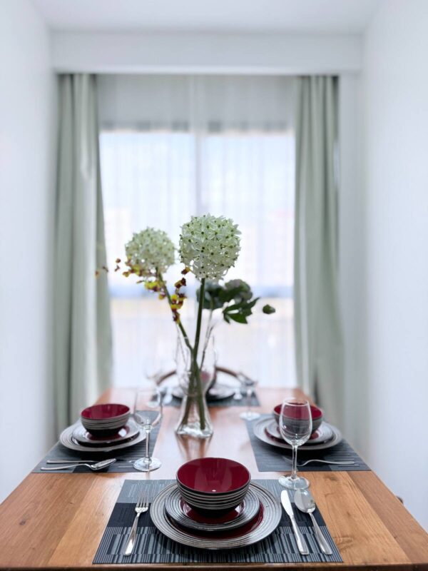 Elegant dining table set with red bowls, wine glasses, and a central vase of white flowers, against sheer curtained windows.
