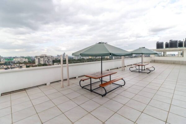 Rooftop terrace with two wooden picnic tables, green umbrellas, and a city skyline view under a cloudy sky.