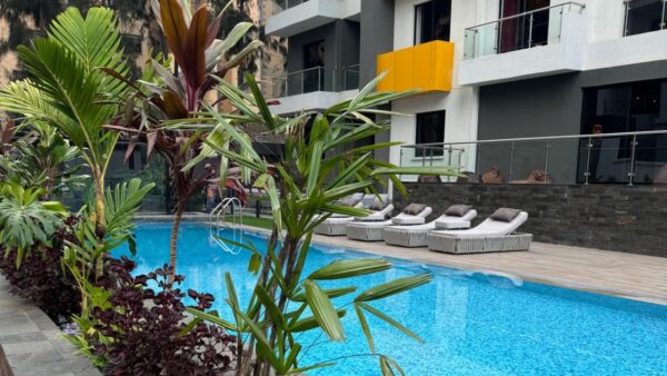 Modern poolside with tropical plants, wicker loungers, and a building with glass balconies and a vibrant yellow panel.