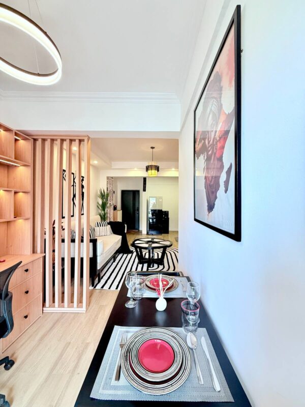 Modern open-concept room with a dining area, decorative wall art, and wooden decor elements featuring a red accent plate setting.