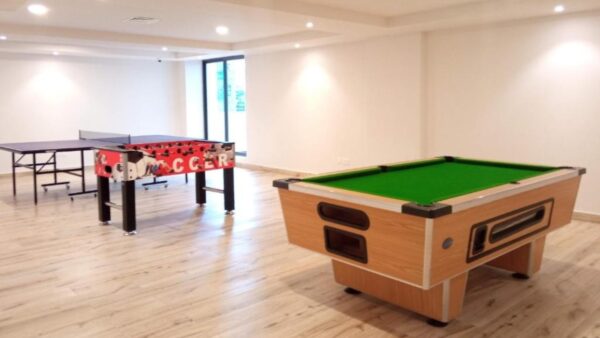 Recreation room with a pool table, foosball table, and ping pong table on wooden floor, bright lighting.