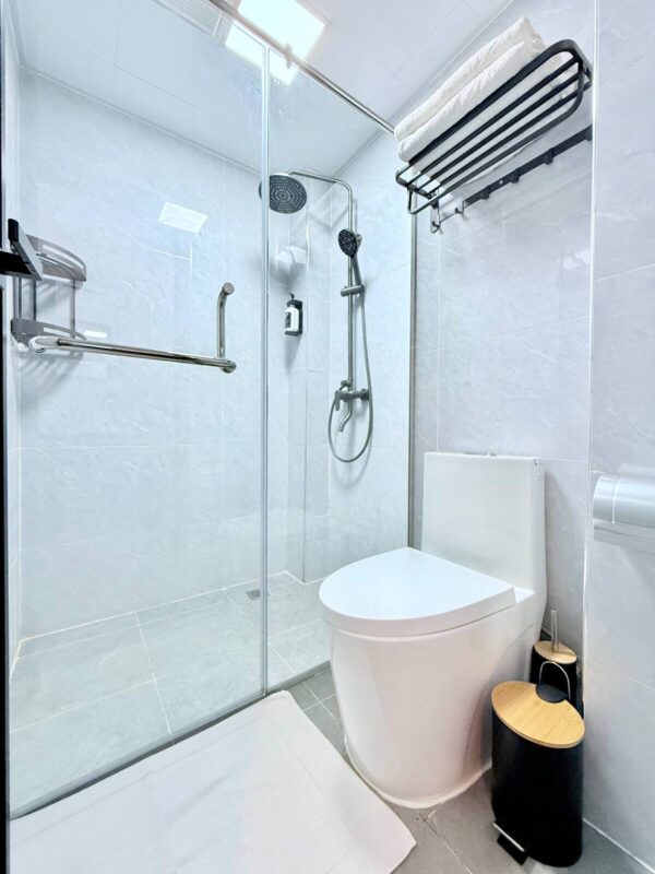 Modern bathroom with glass shower, wall-mounted showerhead, toilet, towel rack, and black trash can on tiled floor.