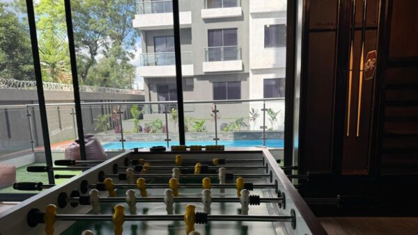 Foosball table indoors, overlooking a pool and modern apartment building through large glass windows.