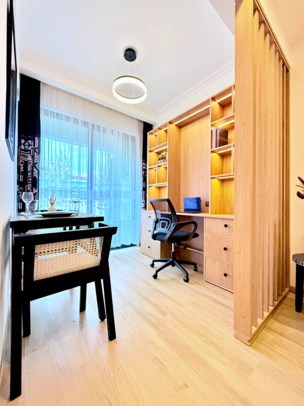 Modern home office with wood desk, shelves, swivel chair, and pendant light. Large window with sheer curtains.