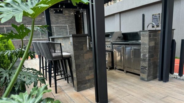 Outdoor kitchen with stone bar, high chairs, stainless steel grill, and lush green plants in the foreground.
