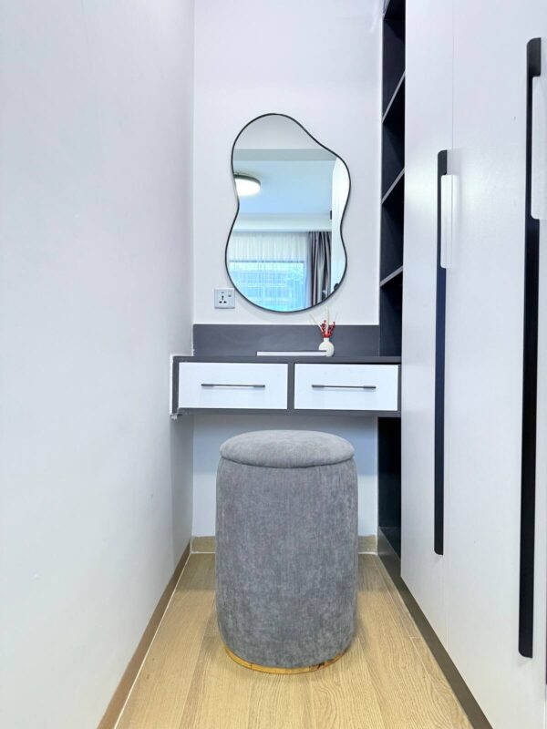Gray stool in front of a vanity with a wavy mirror, white drawers, and a small vase on a wood floor.