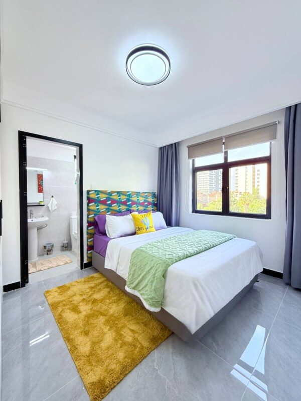 Modern bedroom with a colorful headboard, neatly made bed, green and white bedding, yellow rug, and large window.
