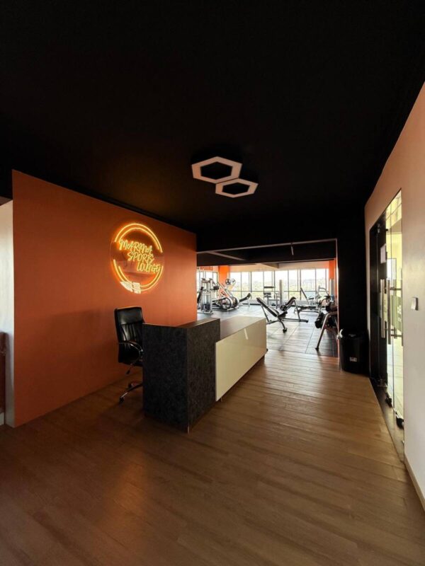Modern gym reception with a desk, swivel chair, neon sign on orange wall, and exercise equipment in the background.