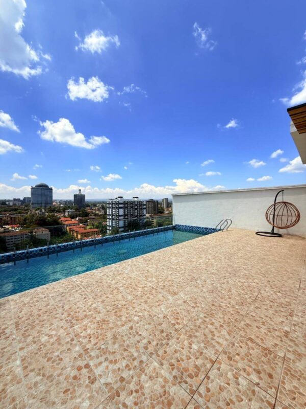 Rooftop infinity pool overlooking a cityscape, with blue sky and scattered clouds above a tan tiled deck.