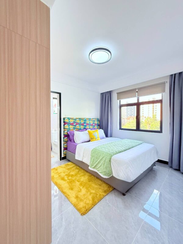 Bright bedroom with colorful headboard, purple and green bedding, yellow rug, modern ceiling light, and window with city view.