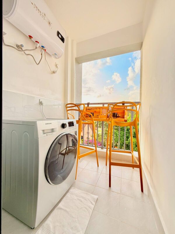 Small balcony with washing machine, yellow chairs, wooden table, water heater, and a view of a clear sky.