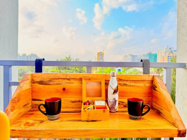 Wooden balcony table with two black mugs, a decorated bottle, and a wooden box. Cityscape and blue sky background.
