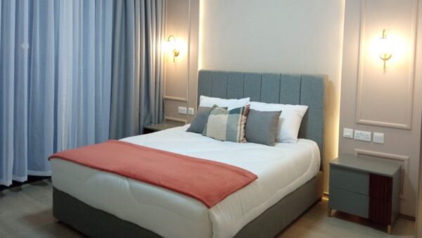 Elegant bedroom with a gray upholstered bed, white and coral linens, pillows, nightstands, wall sconces, and blue curtains.