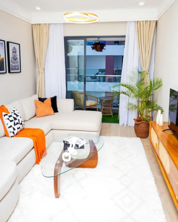 Modern living room with a beige sectional sofa, orange accents, glass coffee table, potted plant, and sliding glass doors.