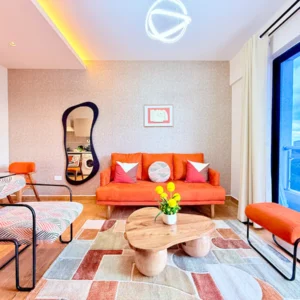Modern living room with orange sofa, abstract mirror, geometric rug, and balcony view. Vibrant decor and curved furniture.