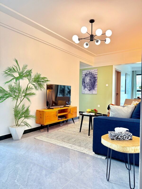 Modern living room with a blue sofa, wooden TV stand, large plant, geometric rug, and a six-light chandelier.