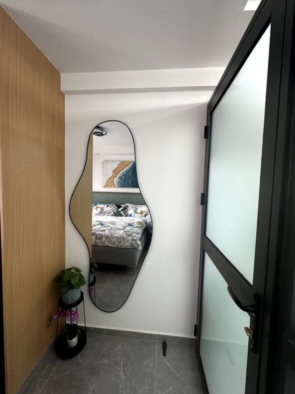 Curved mirror on a white wall reflects a bed with patterned bedding. Potted plants on a small stand beneath.