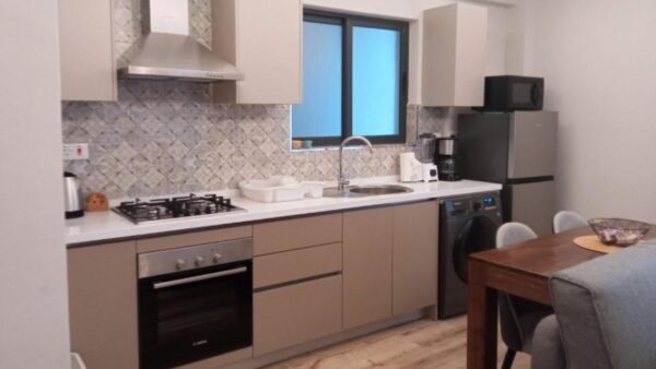 Modern kitchen with beige cabinets, a gas stove, oven, sink, washing machine, fridge, microwave, and dining table with chairs.