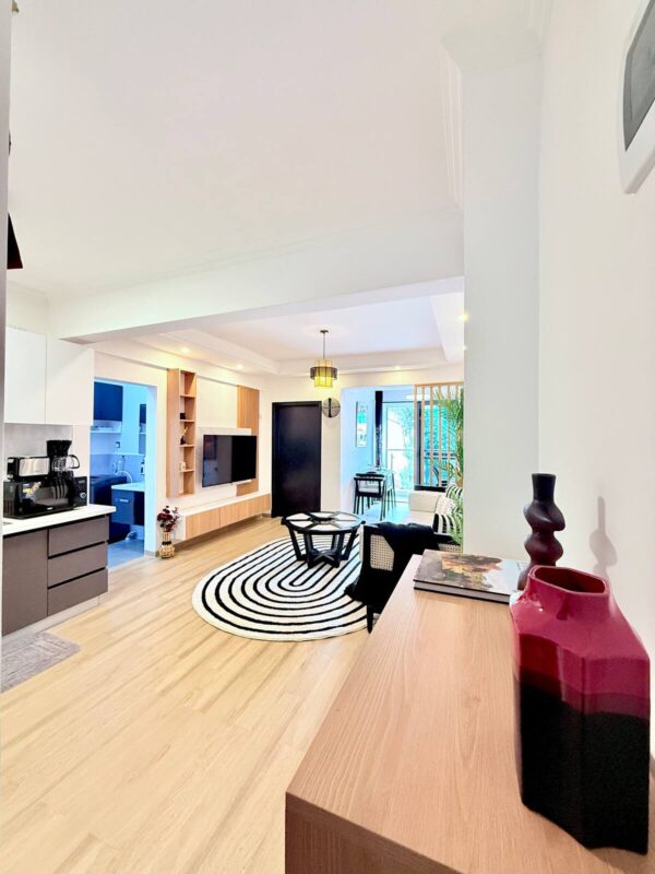 Modern living room with wooden floor, circular striped rug, wall-mounted TV, and a kitchenette on the left.