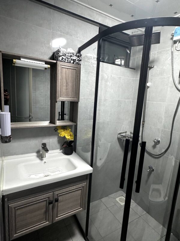 Modern bathroom with a glass shower, gray tiles, wooden vanity with sink, mirror, storage cabinet, and yellow flowers.
