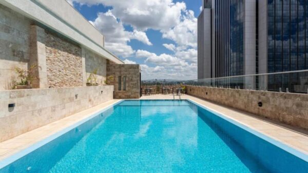 Rooftop pool with clear blue water, surrounded by stone walls, under a partly cloudy sky with a modern building nearby.