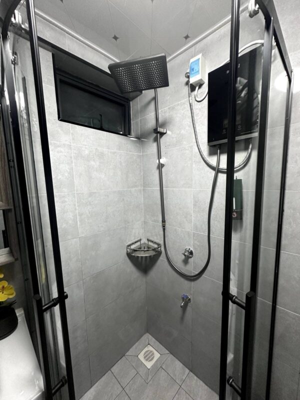 Modern shower with square rainfall head, gray tiled walls, glass doors, corner shelf, and black fixtures under a small window.