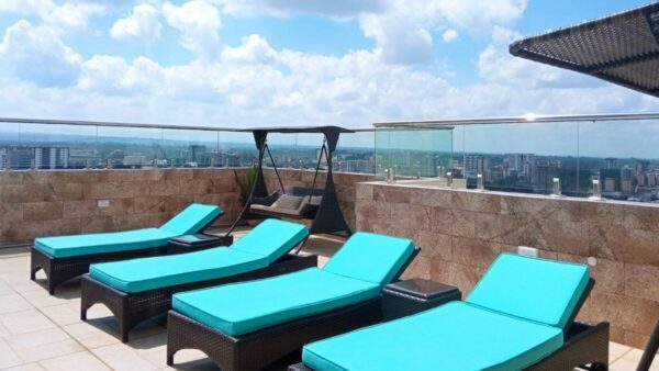 Rooftop terrace with turquoise loungers, cushioned swing, glass railings, cityscape view, and partly cloudy sky.