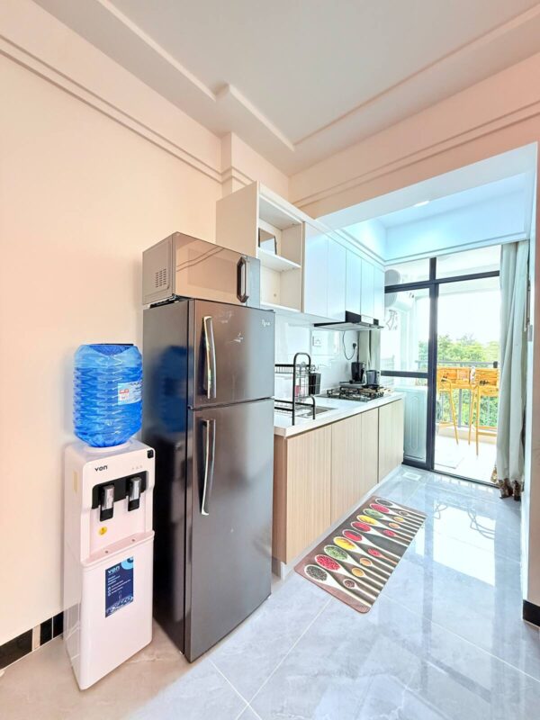 Modern kitchen with a black fridge, microwave, water dispenser, wooden cabinets, and a view of an outdoor balcony.