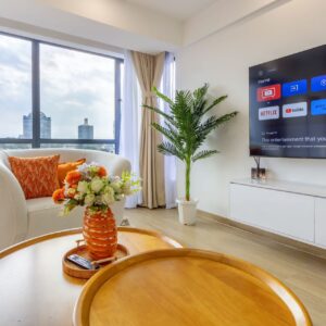 Bright living room with a large window view, white armchair with orange cushions, round wooden coffee table with flowers, potted plant, and wall-mounted TV displaying streaming apps.