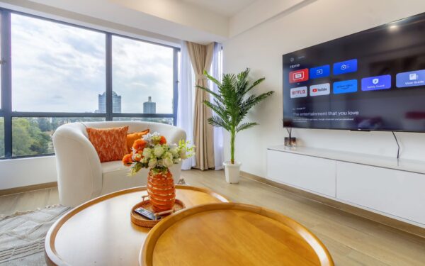 Bright living room with a large window view, white armchair with orange cushions, round wooden coffee table with flowers, potted plant, and wall-mounted TV displaying streaming apps.