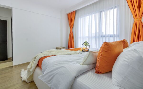 Bright bedroom with white and orange decor, large window with sheer curtains, and a bed adorned with orange pillows and a throw.