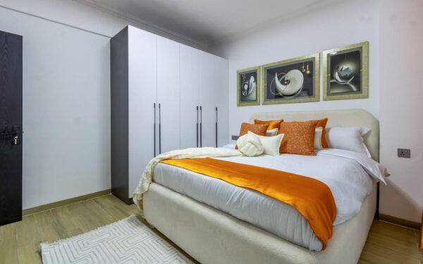 Modern bedroom with white wardrobe, double bed adorned with orange pillows and runner, abstract art on walls.