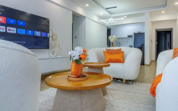 Living room with white armchairs, orange cushions, two round wooden coffee tables with flowers, and a wall-mounted TV visible on the left.