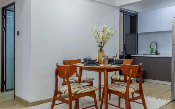 Wooden dining table with four chairs, set with black tableware and a floral centerpiece, adjacent to a modern kitchen.
