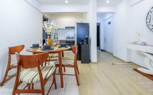Dining area with wooden chairs, striped cushions, set table with dishware, vase of white flowers, and view into the kitchen.