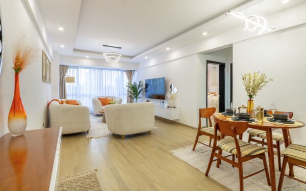 Modern living and dining area with white sofas, a sleek TV, circular dining table, and warm lighting, accented by orange decor.