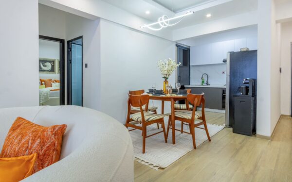 Modern open-plan kitchen and dining area with wooden furniture, a white rug, and contemporary lighting. Orange accents throughout.